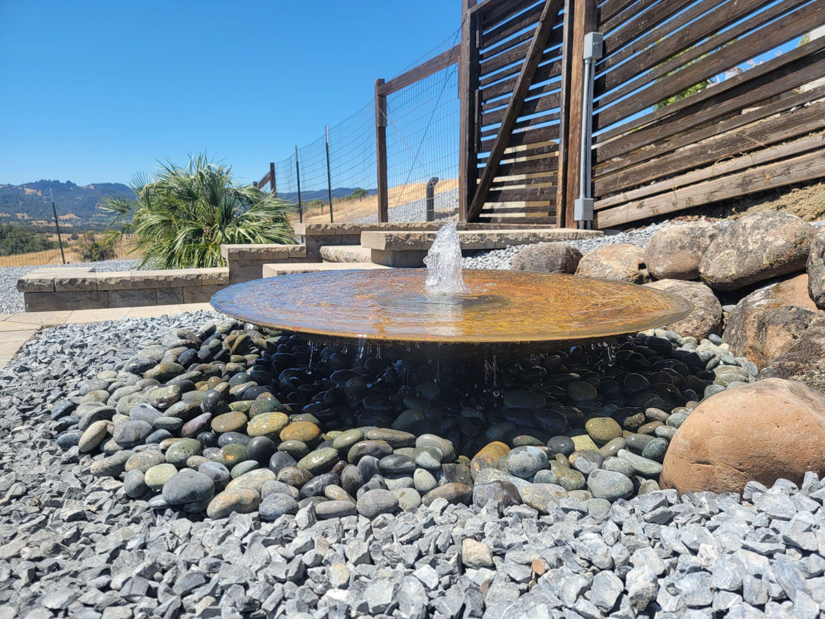 A Water feature sat on pebbles with a fence and steps in the background. 